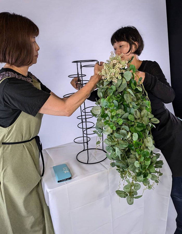 女性二人がフェイクグリーンのフラワーアレンジをスタンドに取り付けている様子。室内で丁寧に装飾中。