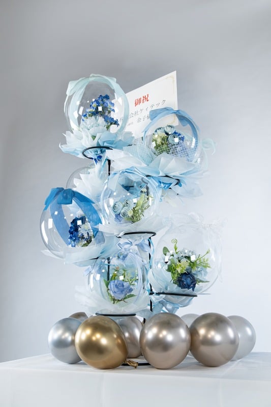 ブルー系のバルーンフラワータワー。爽やかな青と白の花が透明バルーンに包まれ、ゴールドとシルバーの土台に配置されています